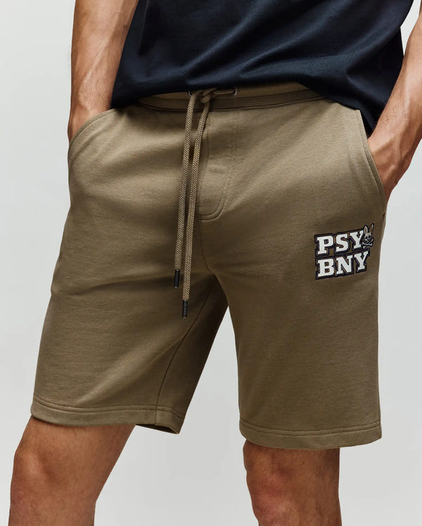 A person in Psycho Bunny's MENS WADE SWEATSHORT - MB0900028, olive green cotton-blend shorts with a "PSY BNY" patch on the left thigh, and a black tee stands in classic athletic style with one hand in the pocket.
