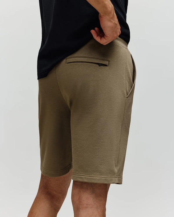 A person wearing Psycho Bunny MENS WADE SWEATSHORT - MB0900028 in olive green, classic athletic style, and a black shirt, shown waist down from the back. The premium cotton blend shorts feature side pockets and a single rear zip pocket.