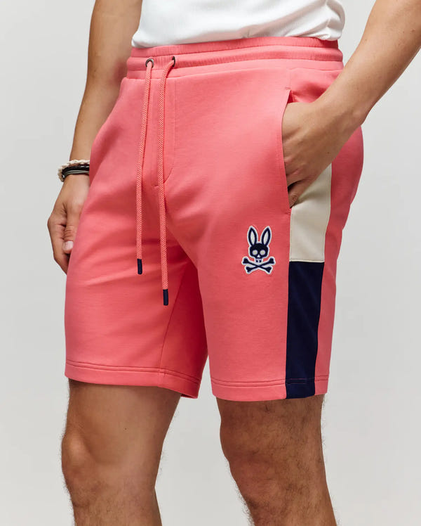 A person wears Psycho Bunny’s MEN'S DAWSON TRACK SHORT - MB0900032 in coral pink with navy and white side stripes, a drawstring waist, and bunny skull logo, styled with a white shirt, bracelets, and one hand in their pocket.
