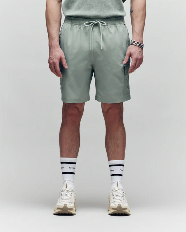 A person faces forward, wearing Psycho Bunny MENS RANDY BOARD SWIM SHORT - MB1000002 in sage green with cargo pockets, paired with a matching shirt, white sneakers, and white socks with black stripes and text; shown from shoulders down on a plain background.