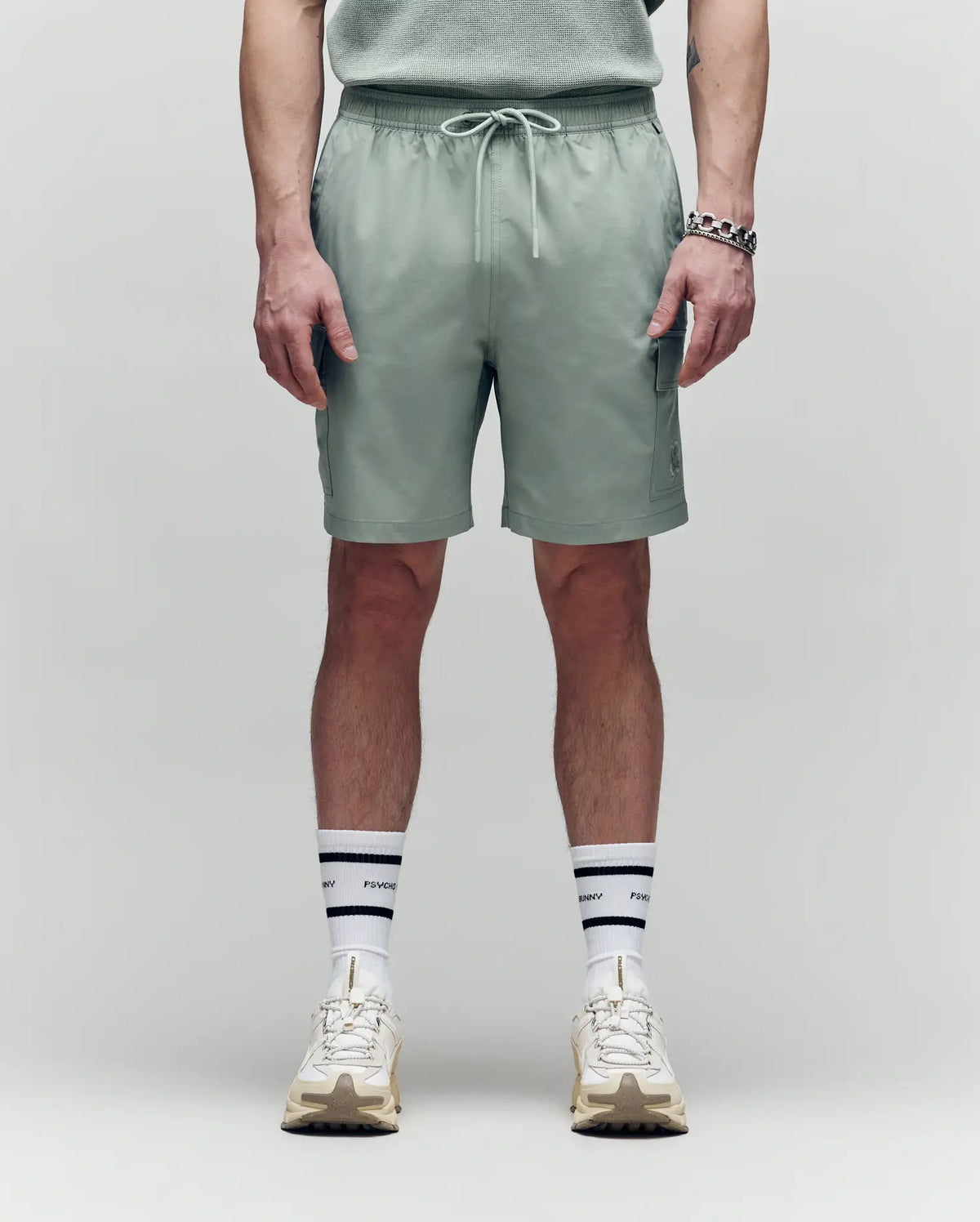 A person faces forward, wearing Psycho Bunny MENS RANDY BOARD SWIM SHORT - MB1000002 in sage green with cargo pockets, paired with a matching shirt, white sneakers, and white socks with black stripes and text; shown from shoulders down on a plain background.