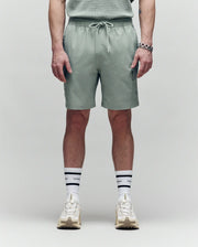 A person faces forward, wearing Psycho Bunny MENS RANDY BOARD SWIM SHORT - MB1000002 in sage green with cargo pockets, paired with a matching shirt, white sneakers, and white socks with black stripes and text; shown from shoulders down on a plain background.