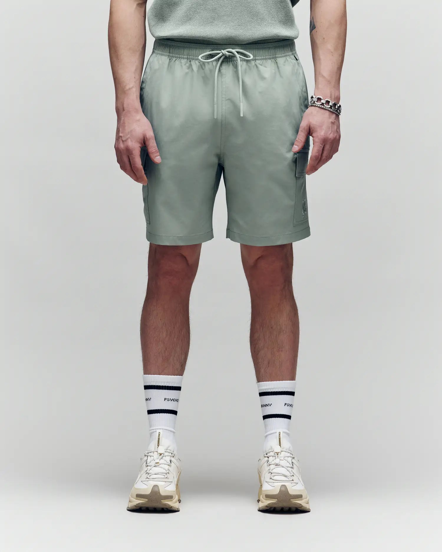 A person faces forward, wearing Psycho Bunny MENS RANDY BOARD SWIM SHORT - MB1000002 in sage green with cargo pockets, paired with a matching shirt, white sneakers, and white socks with black stripes and text; shown from shoulders down on a plain background.