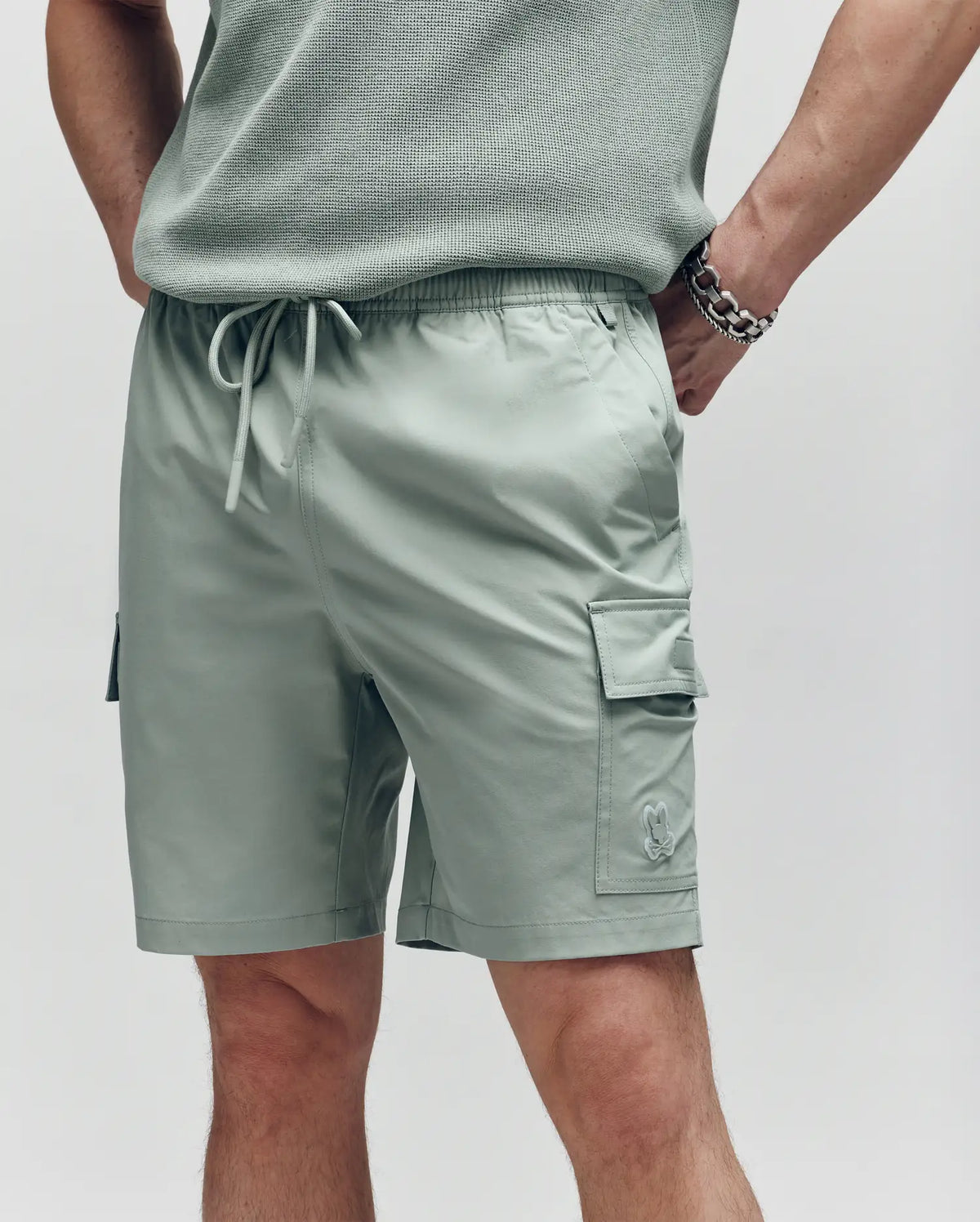 A person wears Psycho Bunny MENS RANDY BOARD SWIM SHORT - MB1000002 in light sage green with cargo pockets and a drawstring waist. Only the lower torso and upper legs are visible, one hand in a pocket, the other arm with a chain bracelet.