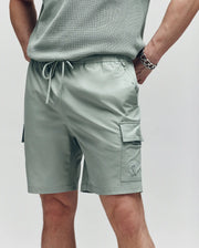 A person wears Psycho Bunny MENS RANDY BOARD SWIM SHORT - MB1000002 in light sage green with cargo pockets and a drawstring waist. Only the lower torso and upper legs are visible, one hand in a pocket, the other arm with a chain bracelet.