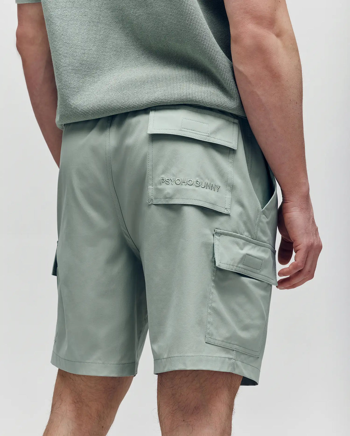 A person in light green Psycho Bunny MENS RANDY BOARD SWIM SHORT - MB1000002 with cargo pockets and "PSYCHO BUNNY" on the back pocket stands facing away, part of a matching textured shirt visible—perfect for casual beachwear.