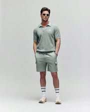 A man wears Psycho Bunny's MENS RANDY BOARD SWIM SHORT - MB1000002 in light green, paired with a matching polo shirt, sunglasses, white socks with black stripes, and white sneakers, standing hands-in-pockets before a plain light background.