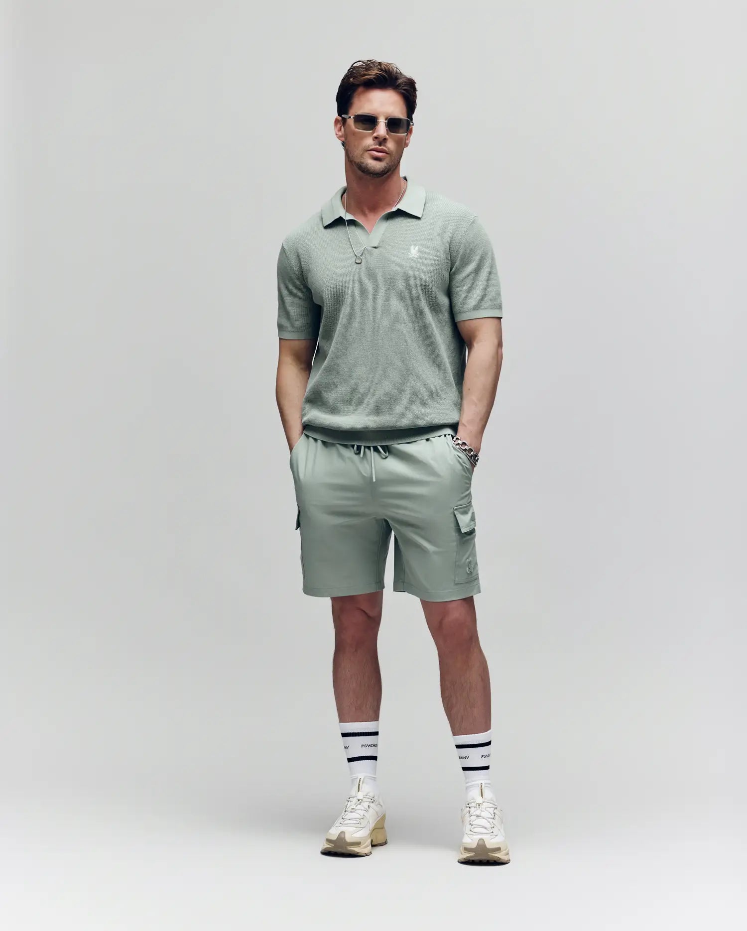 A man wears Psycho Bunny's MENS RANDY BOARD SWIM SHORT - MB1000002 in light green, paired with a matching polo shirt, sunglasses, white socks with black stripes, and white sneakers, standing hands-in-pockets before a plain light background.