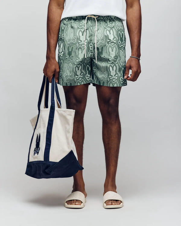 A person wears Psycho Bunny MENS CHUCK ALL OVER PRINT LIGHTWEIGHT SWIM TRUNK - MB1000003, a white T-shirt, and white slide sandals, holding a cream and navy tote with a small black graphic against a plain background.