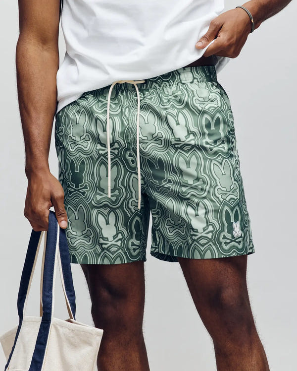 A man wears a white tee and Psycho Bunny MENS CHUCK ALL OVER PRINT LIGHTWEIGHT SWIM TRUNK (MB1000003) in green, with abstract bunny shapes, holding a beige tote with dark handles against a plain light background.