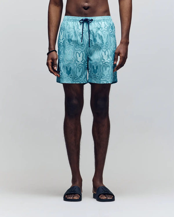 A person wears Psycho Bunny MENS CHUCK ALL OVER PRINT LIGHTWEIGHT SWIM TRUNK - MB1000003 in blue, paired with black slide sandals and a bracelet. The lower half of their body stands against a plain light background.