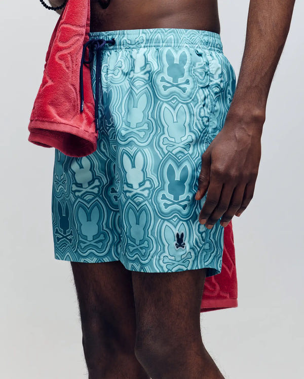 A person is seen from torso to upper legs wearing Psycho Bunny’s MENS CHUCK ALL OVER PRINT LIGHTWEIGHT SWIM TRUNK (MB1000003) in light blue with an abstract bunny pattern, made from recycled polyester. A red towel is draped over their shoulder.