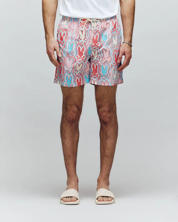 A person in a white t-shirt, Psycho Bunny MENS CHUCK ALL OVER PRINT LIGHTWEIGHT SWIM TRUNK - MB1000003 with red, blue, and pink designs, white slide sandals, and a thin bracelet stands against a plain light background.