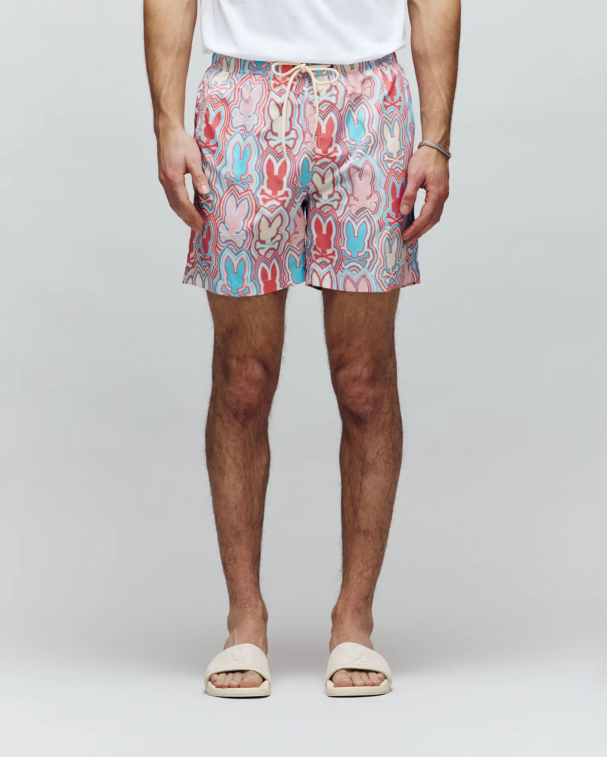 A person in a white t-shirt, Psycho Bunny MENS CHUCK ALL OVER PRINT LIGHTWEIGHT SWIM TRUNK - MB1000003 with red, blue, and pink designs, white slide sandals, and a thin bracelet stands against a plain light background.