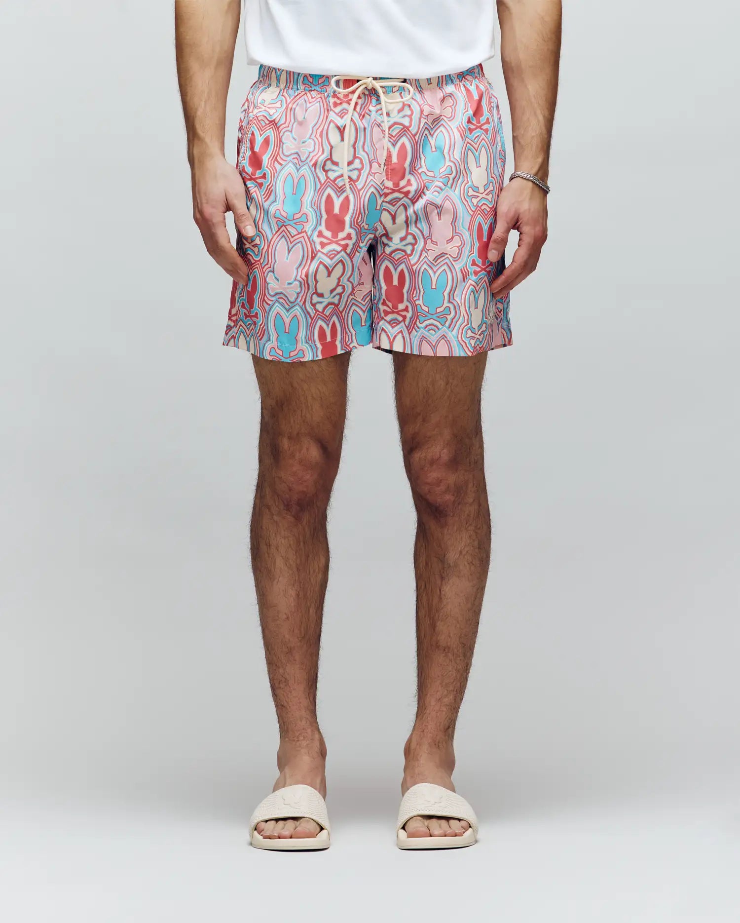 A person in a white t-shirt, Psycho Bunny MENS CHUCK ALL OVER PRINT LIGHTWEIGHT SWIM TRUNK - MB1000003 with red, blue, and pink designs, white slide sandals, and a thin bracelet stands against a plain light background.