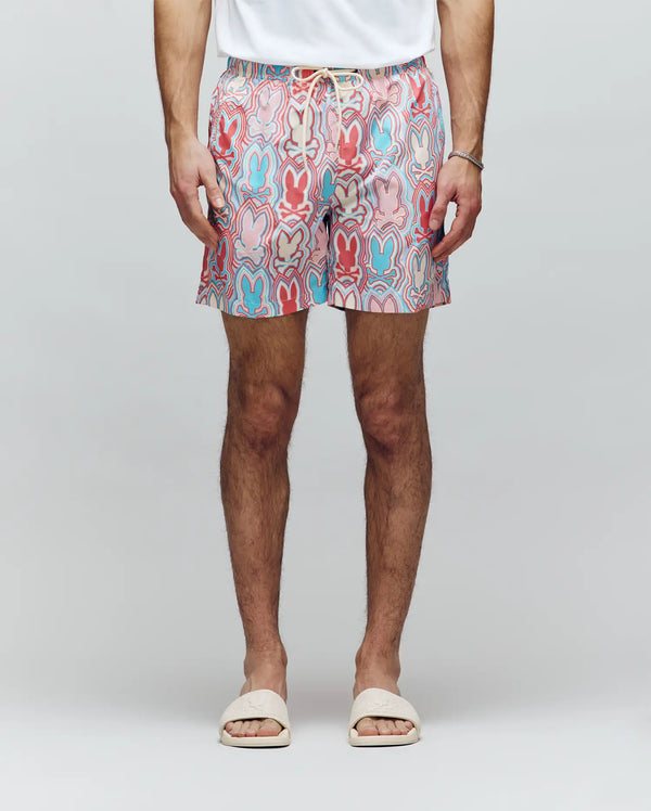 A person in a white t-shirt, Psycho Bunny MENS CHUCK ALL OVER PRINT LIGHTWEIGHT SWIM TRUNK - MB1000003 with red, blue, and pink designs, white slide sandals, and a thin bracelet stands against a plain light background.