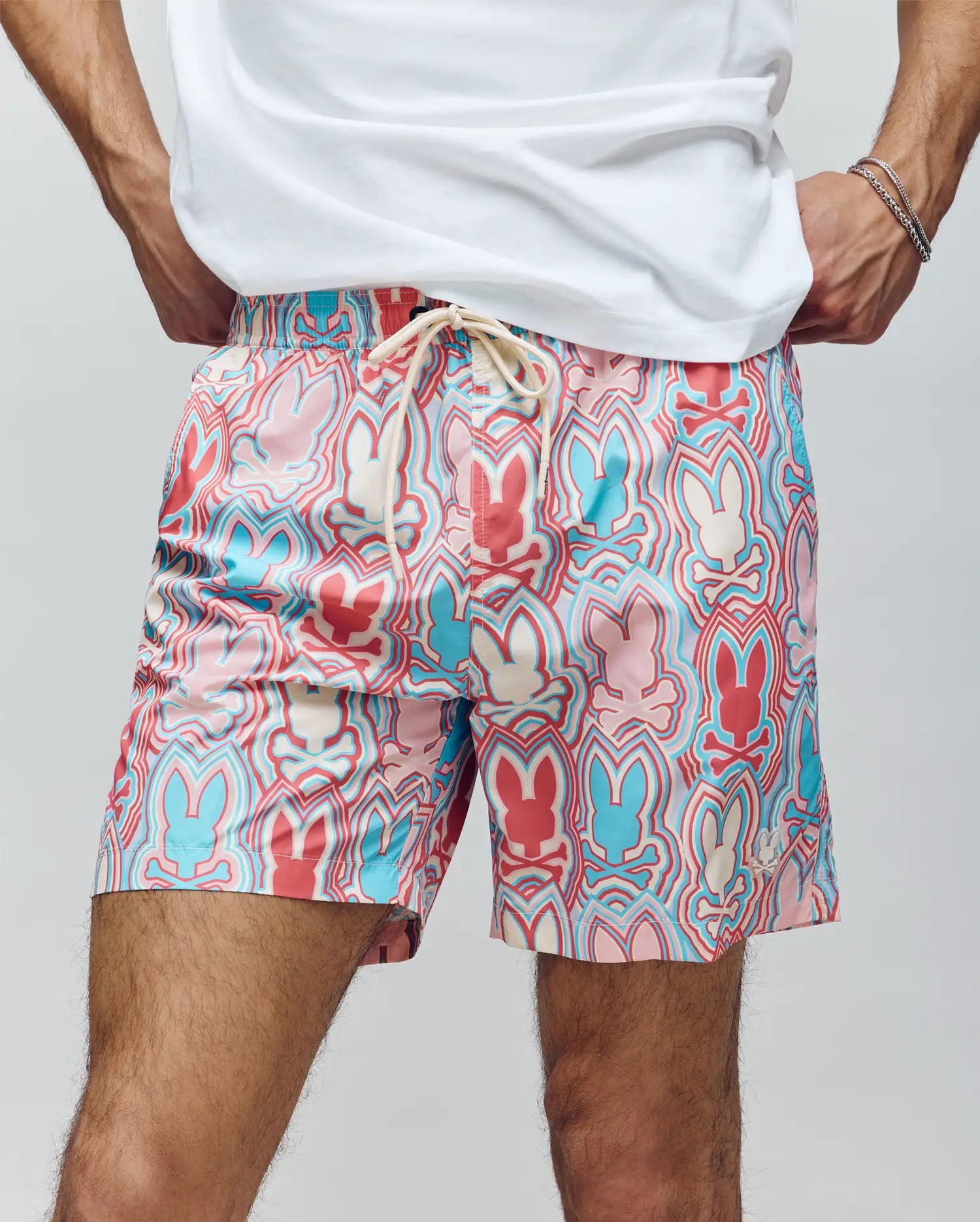 A person wears Psycho Bunny MENS CHUCK ALL OVER PRINT LIGHTWEIGHT SWIM TRUNK - MB1000003, colorful with blue, red, and pink outlined bunny silhouettes and a white drawstring, made from quick dry recycled polyester. They're also in a white shirt and bracelet.