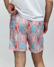 A person stands with one hand on their hip, facing away, wearing Psycho Bunny MENS CHUCK ALL OVER PRINT LIGHTWEIGHT SWIM TRUNK (MB1000003) featuring recycled polyester and a red, blue, and pink outlined bunny pattern.