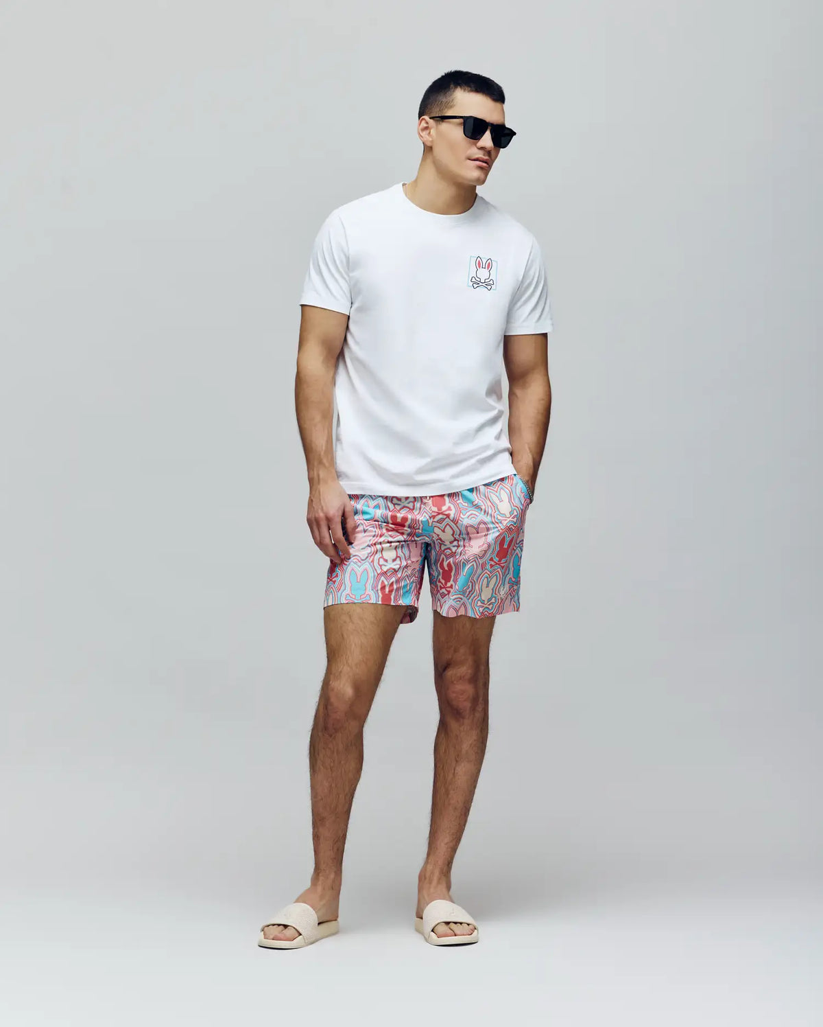 A man in black sunglasses, a white tee with a chest graphic, and Psycho Bunny MENS CHUCK ALL OVER PRINT LIGHTWEIGHT SWIM TRUNK - MB1000003 made from recycled polyester stands with hands in pockets against a plain light background, wearing beige slide sandals.