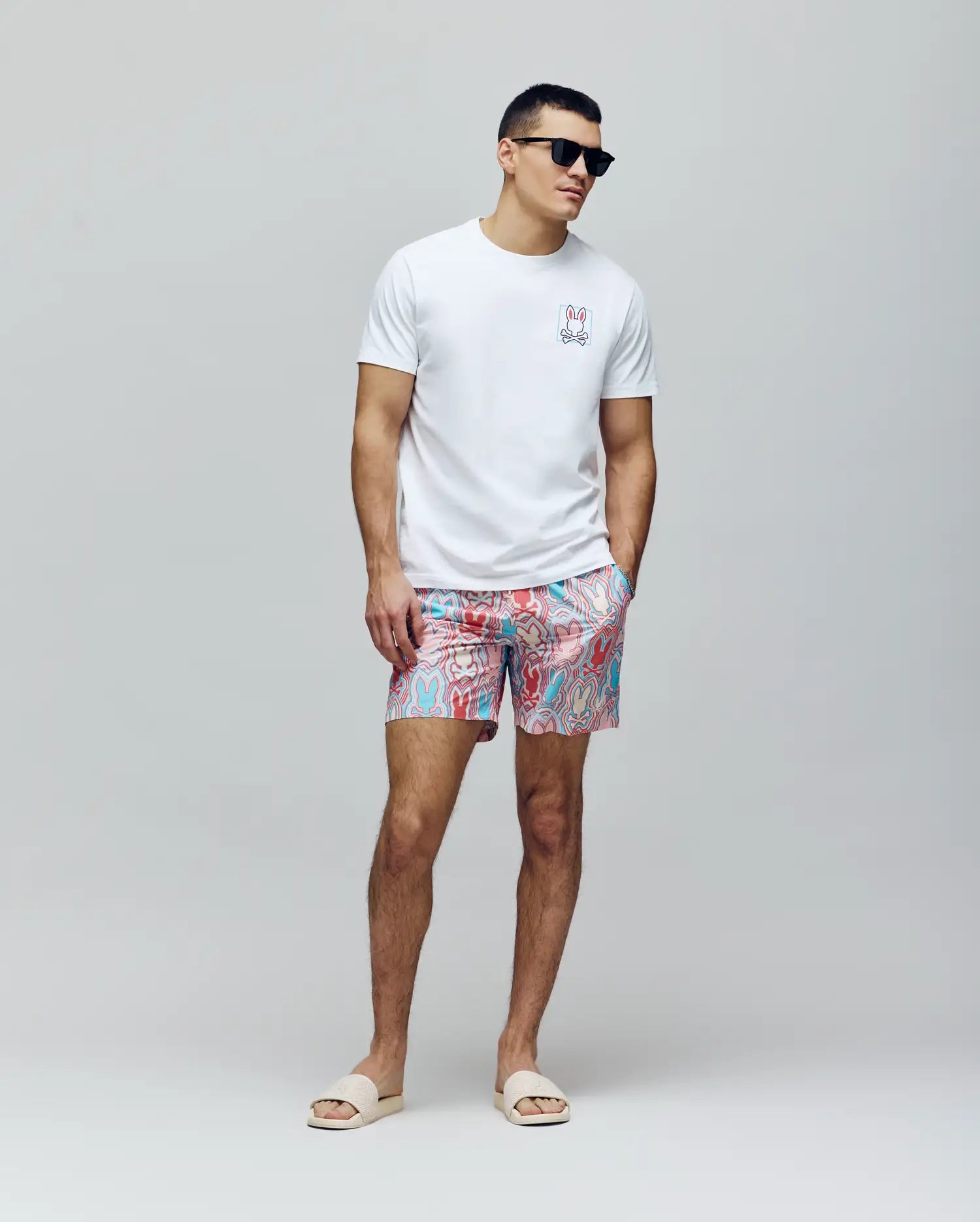 A man in black sunglasses, a white tee with a chest graphic, and Psycho Bunny MENS CHUCK ALL OVER PRINT LIGHTWEIGHT SWIM TRUNK - MB1000003 made from recycled polyester stands with hands in pockets against a plain light background, wearing beige slide sandals.