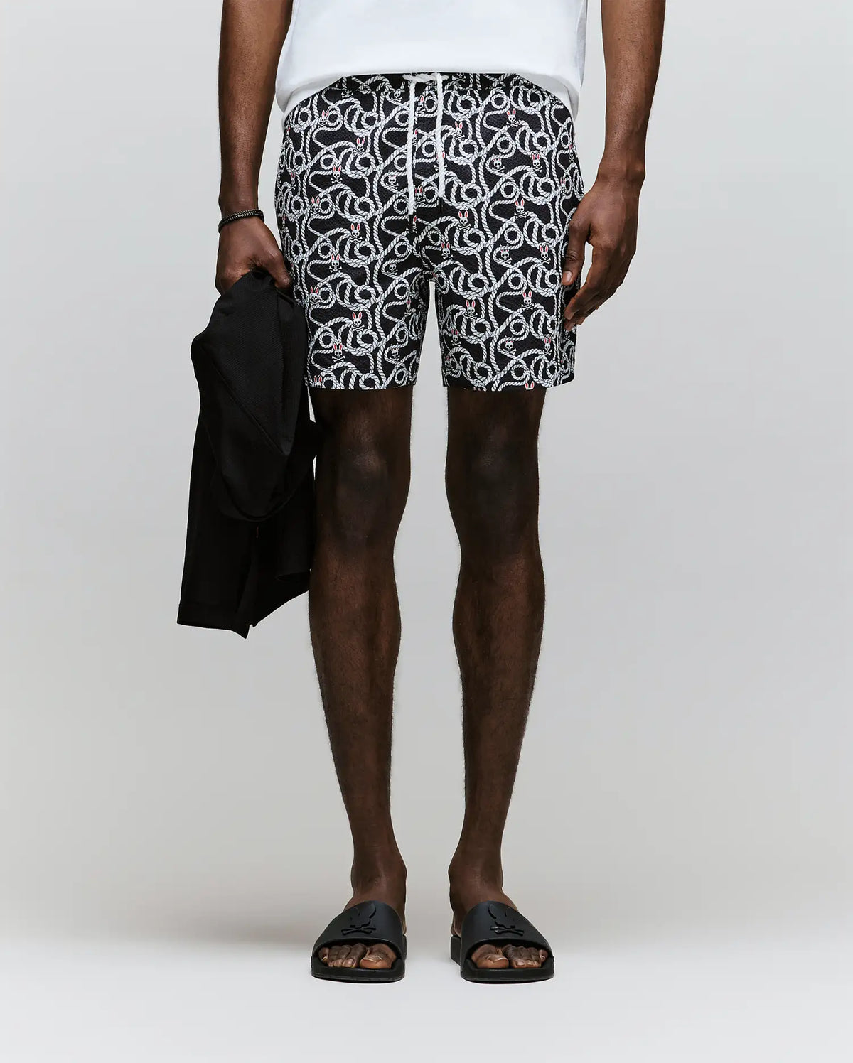 A person stands against a plain background, wearing Psycho Bunny MENS CYRUS ALL OVER PRINT SWIM TRUNK - MB1000004, a white shirt, black slide sandals, and holding a dark jacket in their left hand. The image shows them from shoulders to feet.