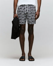A person stands against a plain background, wearing Psycho Bunny MENS CYRUS ALL OVER PRINT SWIM TRUNK - MB1000004, a white shirt, black slide sandals, and holding a dark jacket in their left hand. The image shows them from shoulders to feet.
