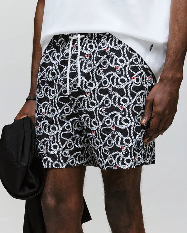 A person is wearing Psycho Bunny's MENS CYRUS ALL OVER PRINT SWIM TRUNK - MB1000004, featuring a black and white rope and knot print, quick-dry seersucker fabric, silicone Bunny patch, white drawstring with red accents, paired with a white shirt.