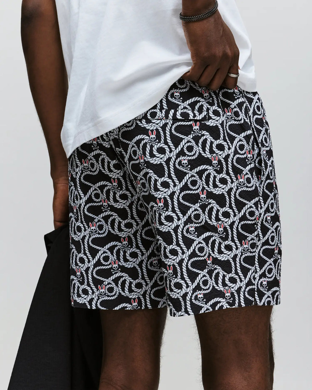 Wearing Psycho Bunny's MENS CYRUS ALL OVER PRINT SWIM TRUNK - MB1000004, a person sports black and white seersucker swim trunks with coiled snake motifs, a white t-shirt, holds a dark garment, and rests their hand on the shorts' waistband.