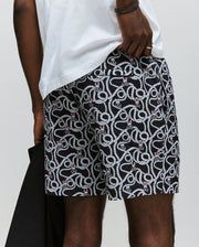 Wearing Psycho Bunny's MENS CYRUS ALL OVER PRINT SWIM TRUNK - MB1000004, a person sports black and white seersucker swim trunks with coiled snake motifs, a white t-shirt, holds a dark garment, and rests their hand on the shorts' waistband.