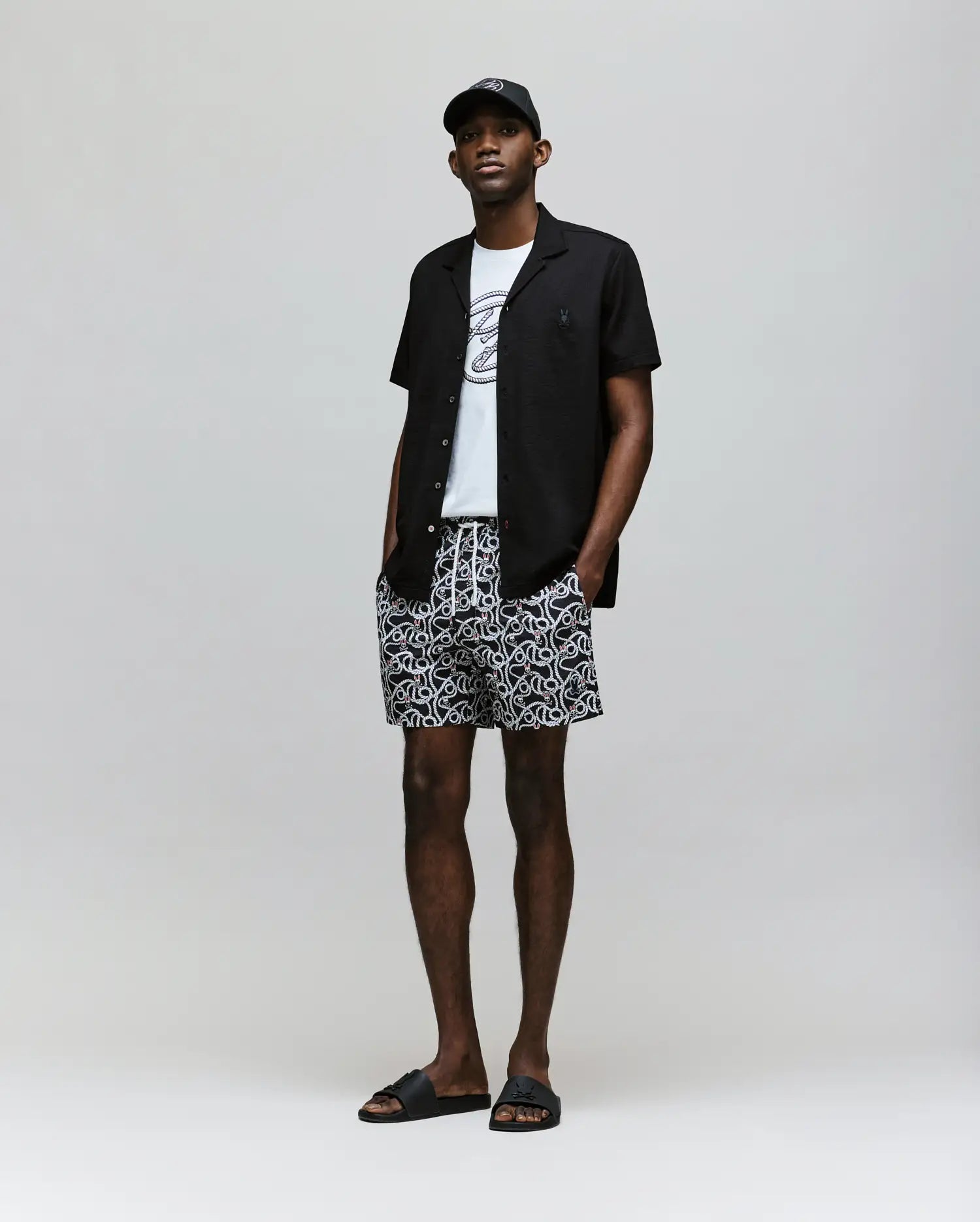 A man stands against a plain background wearing a black cap, open black short-sleeve shirt, white graphic t-shirt, Psycho Bunny MENS CYRUS ALL OVER PRINT SWIM TRUNK (MB1000004), and black slides. His hands are in his pockets.