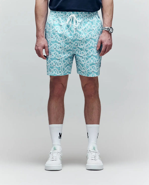 A person sports Psycho Bunny's MENS CYRUS ALL OVER PRINT SWIM TRUNK - MB1000004, a dark shirt, logo white socks, and white sneakers against a light backdrop. Their face is not shown—capturing an effortless coastal cool vibe.