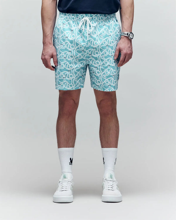 A person sports Psycho Bunny's MENS CYRUS ALL OVER PRINT SWIM TRUNK - MB1000004, a dark shirt, logo white socks, and white sneakers against a light backdrop. Their face is not shown—capturing an effortless coastal cool vibe.