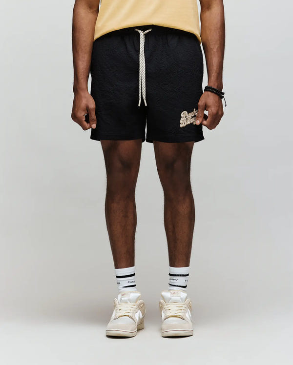 A person in a yellow shirt, white socks, beige sneakers, and classic fit black Psycho Bunny MENS ELIAS SWIM TRUNK (MB1000008) with a white drawstring stands against a plain light gray background.