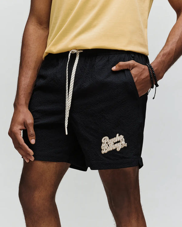 A person wears a yellow t-shirt and Psycho Bunny MENS ELIAS SWIM TRUNK - MB1000008, classic fit in black with a drawstring and logo, standing with one hand in their pocket against a plain background.