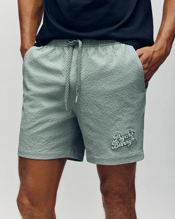 A person wears Psycho Bunny MENS ELIAS SWIM TRUNK - MB1000008, classic fit textured light gray shorts with a drawstring and logo on the left leg, paired with a black t-shirt; hands in pockets, only torso and legs shown.