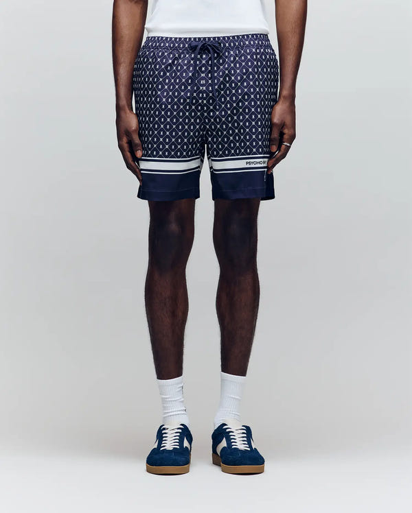 A person stands against a plain light background wearing Psycho Bunny MENS HUNTER ALL OVER PRINT SWIM TRUNK - MB1000011, featuring white and blue striped hems and no-chafe netting, paired with white socks and navy sneakers.