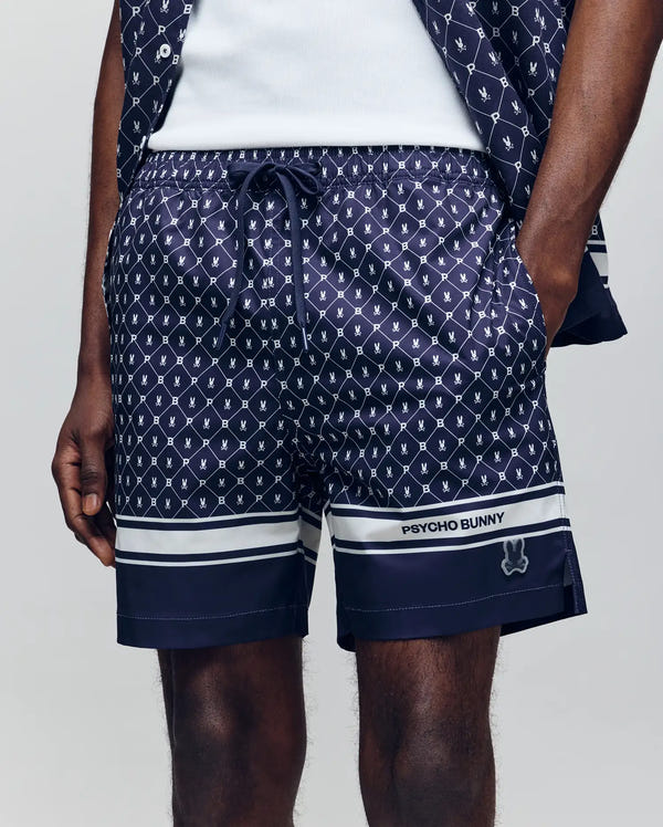 A person wears the Psycho Bunny MENS HUNTER ALL OVER PRINT SWIM TRUNK (MB1000011)—navy shorts with white bunny logos, quick-dry fabric, and "Psycho Bunny" branding on the left leg. Hands are in pockets; a matching shirt is partly visible.