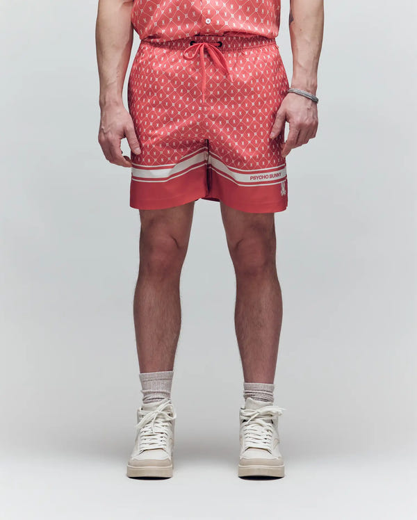 A man faces forward with only his lower body visible, wearing Psycho Bunny’s MENS HUNTER ALL OVER PRINT SWIM TRUNK (MB1000011), light gray socks, and white high-top sneakers against a plain light background.