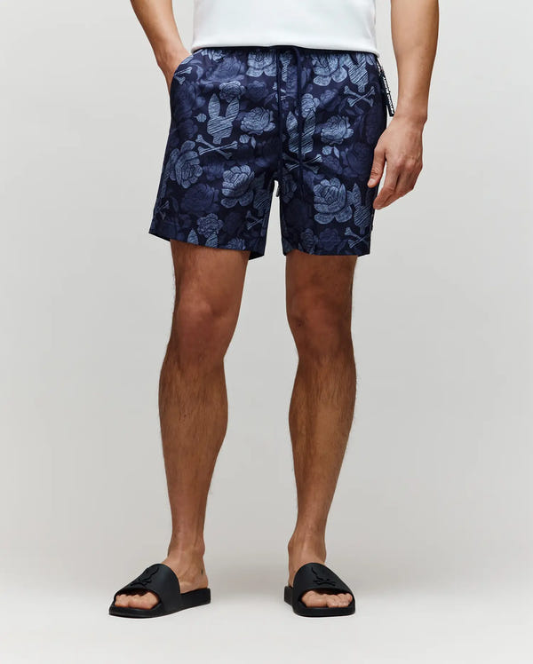 A person wearing Psycho Bunny MENS ROWEN SWIM TRUNK - MB1000023 in navy floral print, a white shirt, and black slide sandals stands against a plain light background. Only the lower half of the body is visible.