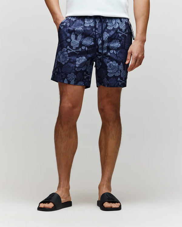 A person wearing Psycho Bunny MENS ROWEN SWIM TRUNK - MB1000023 in navy floral print, a white shirt, and black slide sandals stands against a plain light background. Only the lower half of the body is visible.