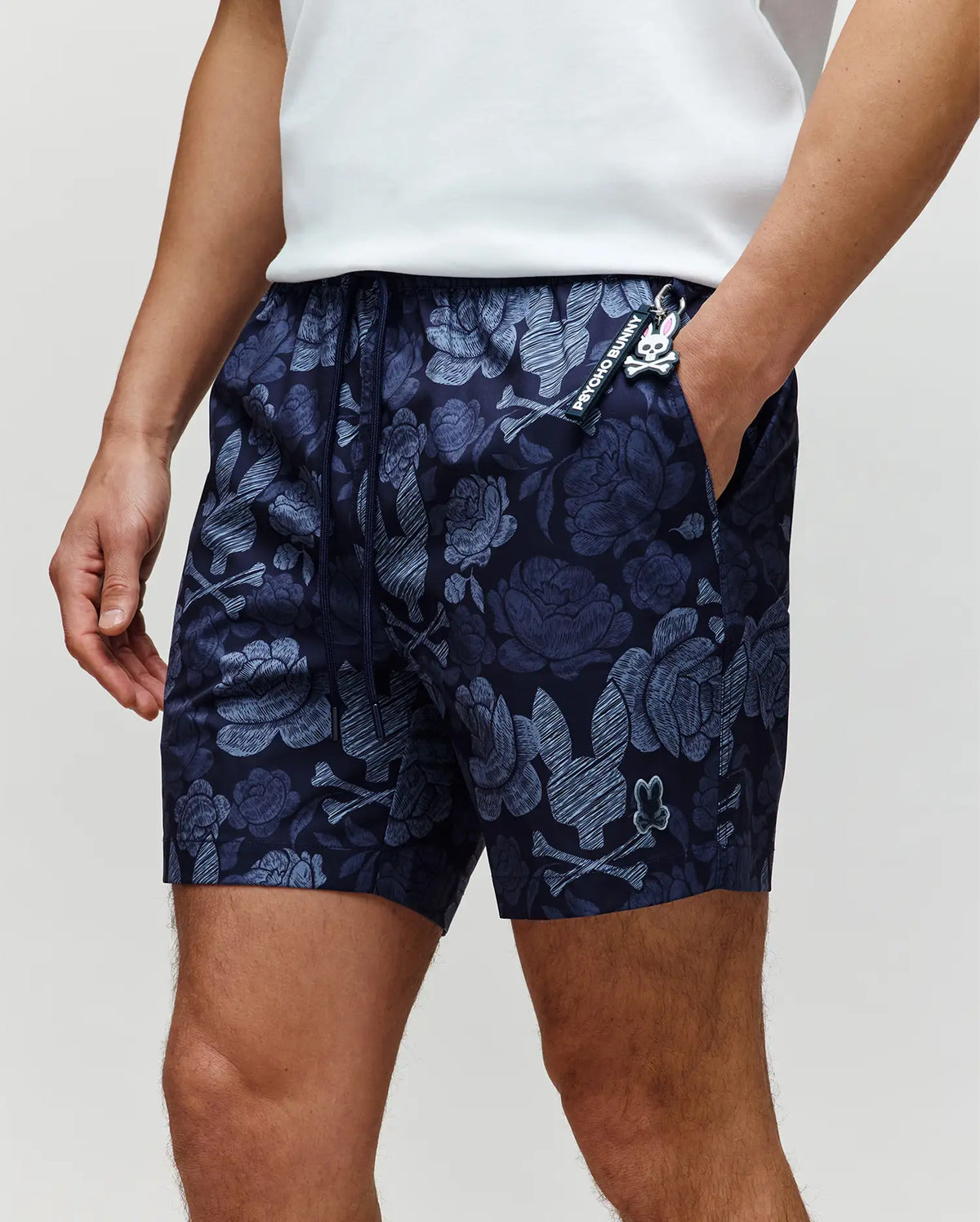 A person wearing a white shirt and Psycho Bunny MENS ROWEN SWIM TRUNK - MB1000023 in dark blue with a floral pattern, hands in pockets, highlights the quick-dry swimwear and upper legs.