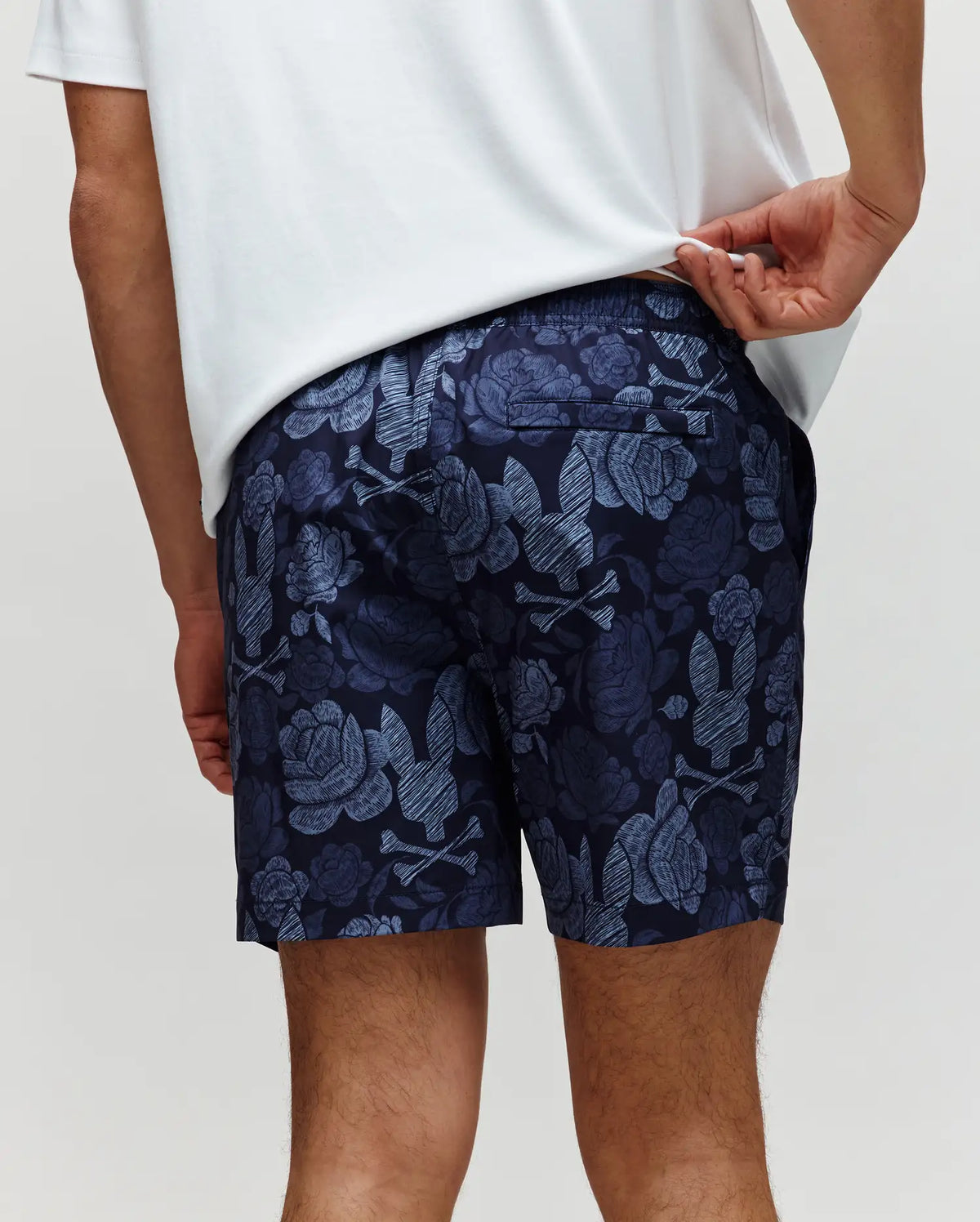 A person in a white shirt lifts it to reveal the stylish Psycho Bunny MENS ROWEN SWIM TRUNK - MB1000023, navy blue with a light blue floral pattern, shown from the back.