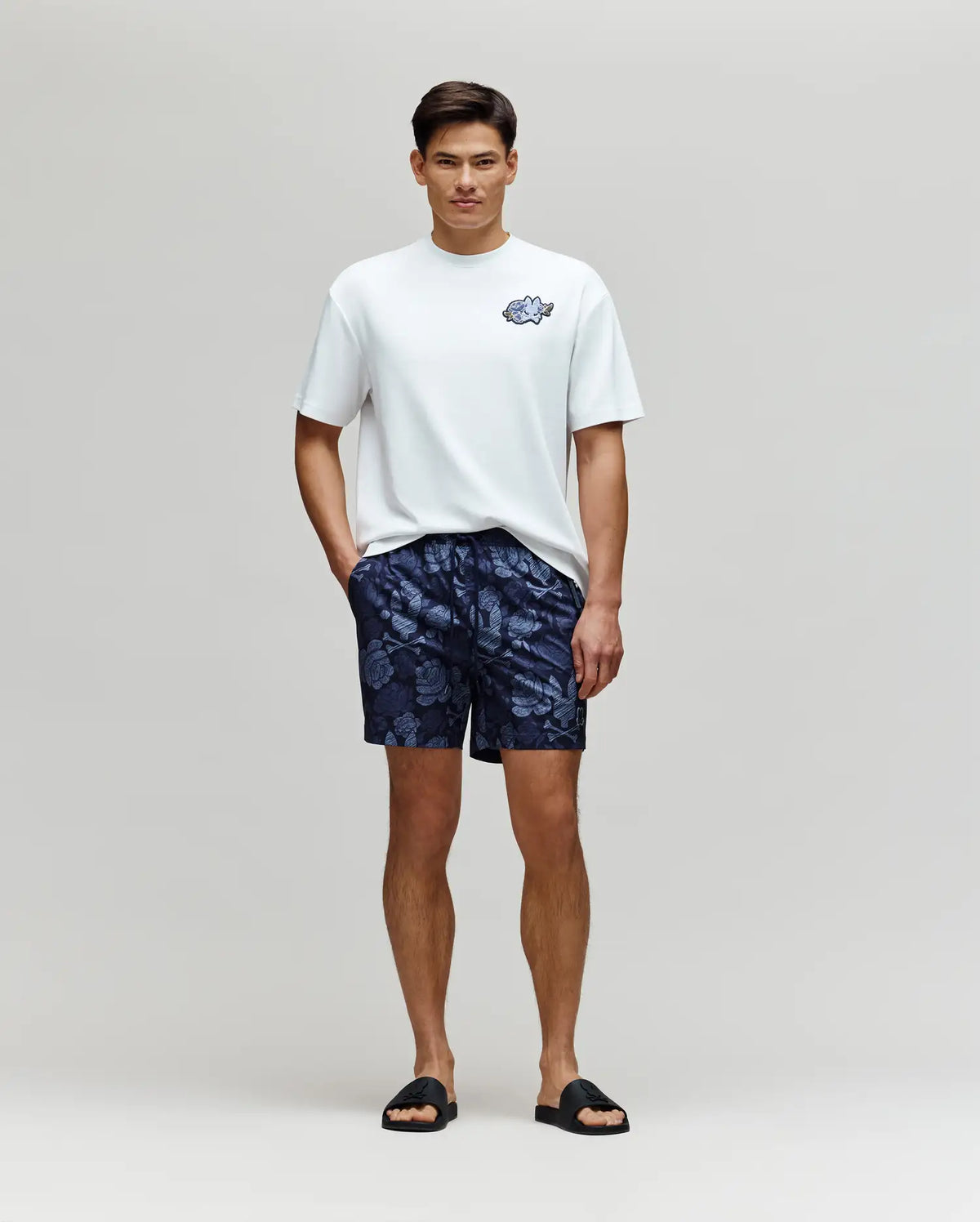 A man stands against a light background wearing a white tee, black slides, and Psycho Bunny MENS ROWEN SWIM TRUNK - MB1000023 in navy blue, with his hands resting in the pockets of the quick-dry swimwear.