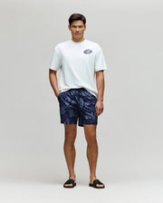 A man stands against a light background wearing a white tee, black slides, and Psycho Bunny MENS ROWEN SWIM TRUNK - MB1000023 in navy blue, with his hands resting in the pockets of the quick-dry swimwear.