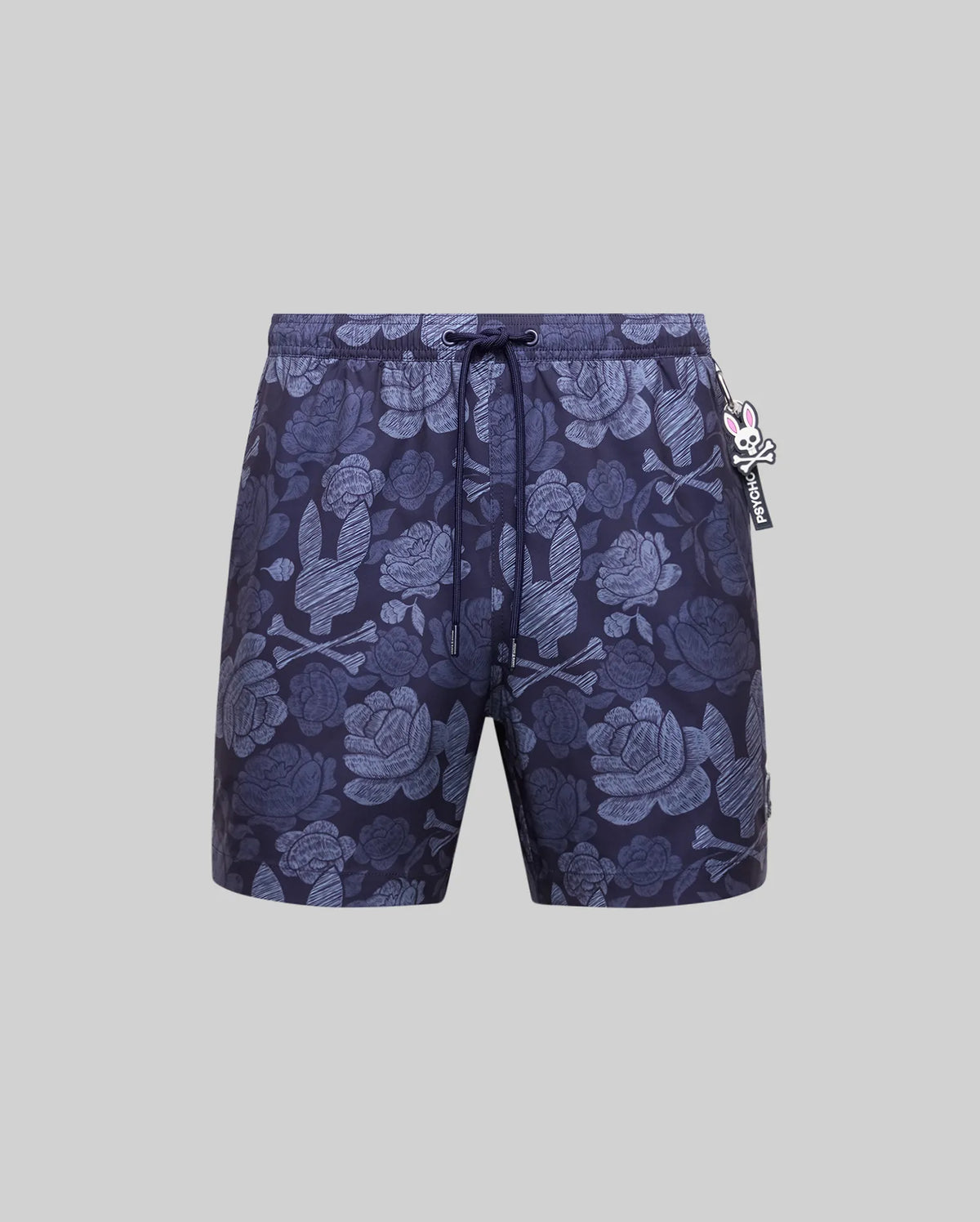 The Psycho Bunny MENS ROWEN SWIM TRUNK (MB1000023) features dark blue floral designs with light blue skulls, a drawstring waistband, and a small rabbit-shaped keychain on the left, shown against a plain gray background.