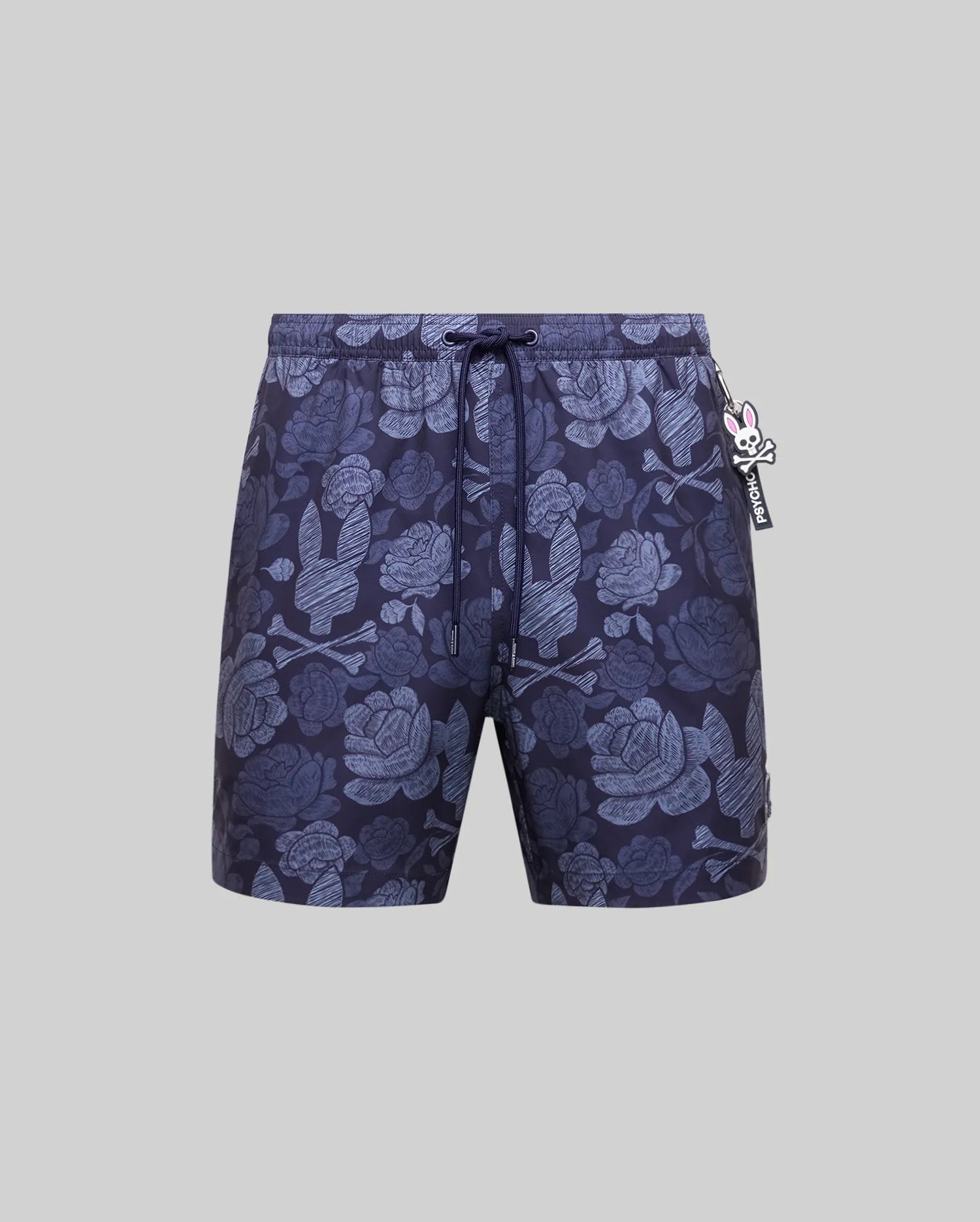 The Psycho Bunny MENS ROWEN SWIM TRUNK (MB1000023) features dark blue floral designs with light blue skulls, a drawstring waistband, and a small rabbit-shaped keychain on the left, shown against a plain gray background.