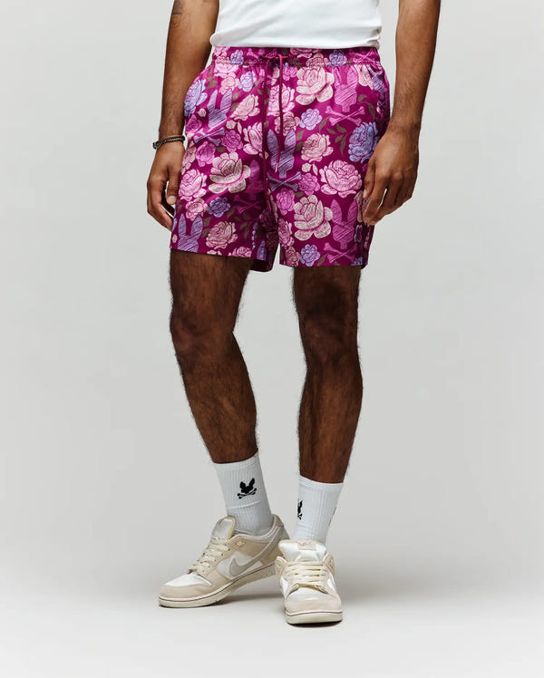 A person models the Psycho Bunny MENS ROWEN SWIM TRUNK - MB1000023, featuring vibrant pink and purple floral print with mesh lining, paired with a white shirt, white crew socks with black designs, and white sneakers against a light gray background.