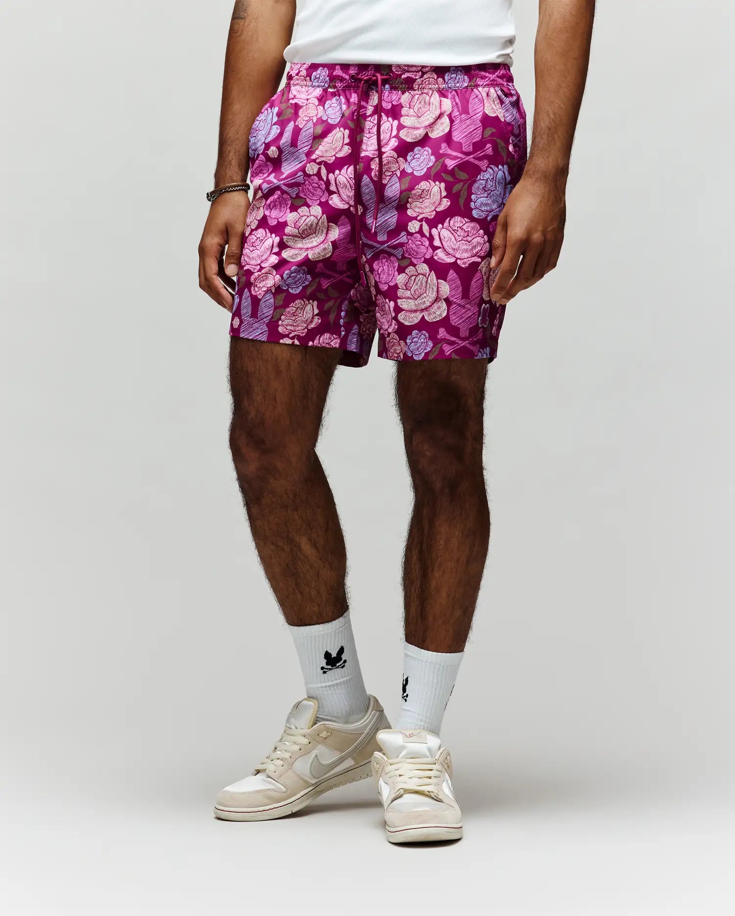 A person models the Psycho Bunny MENS ROWEN SWIM TRUNK - MB1000023, featuring vibrant pink and purple floral print with mesh lining, paired with a white shirt, white crew socks with black designs, and white sneakers against a light gray background.