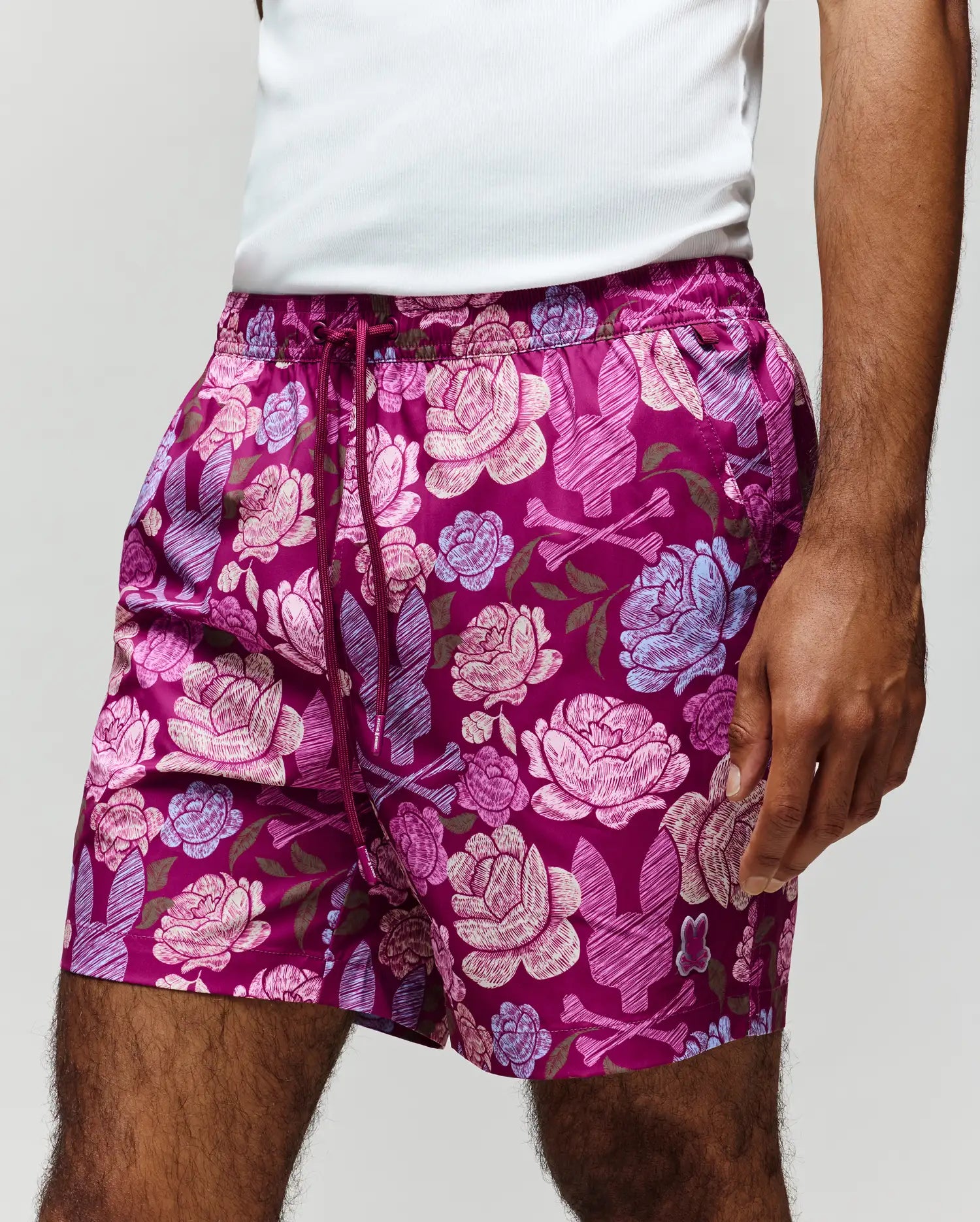 A person wears the Psycho Bunny MENS ROWEN SWIM TRUNK (MB1000023), featuring bold pink, lavender, and magenta floral patterns on quick-dry fabric with mesh lining, shown from lower torso to thighs against a plain background.