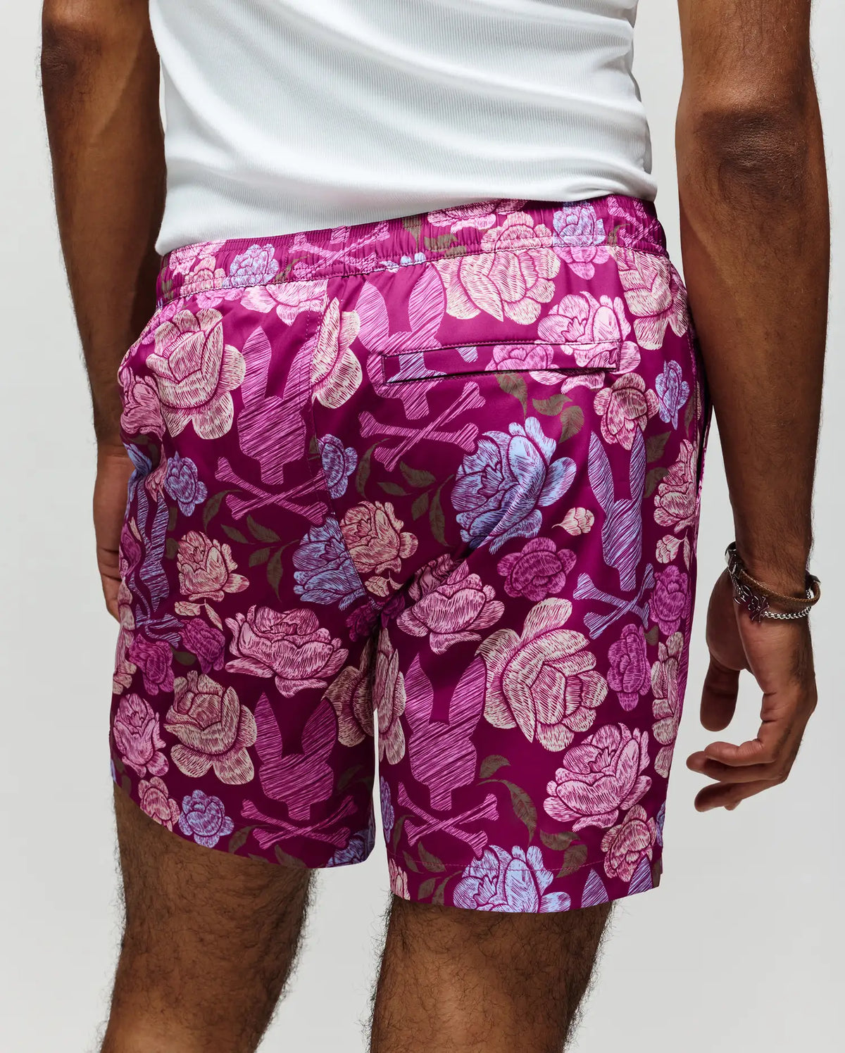 A person stands facing away, wearing Psycho Bunny MENS ROWEN SWIM TRUNK - MB1000023: quick-dry, magenta floral swimwear with bold pink and blue patterns, paired with a white tank top.